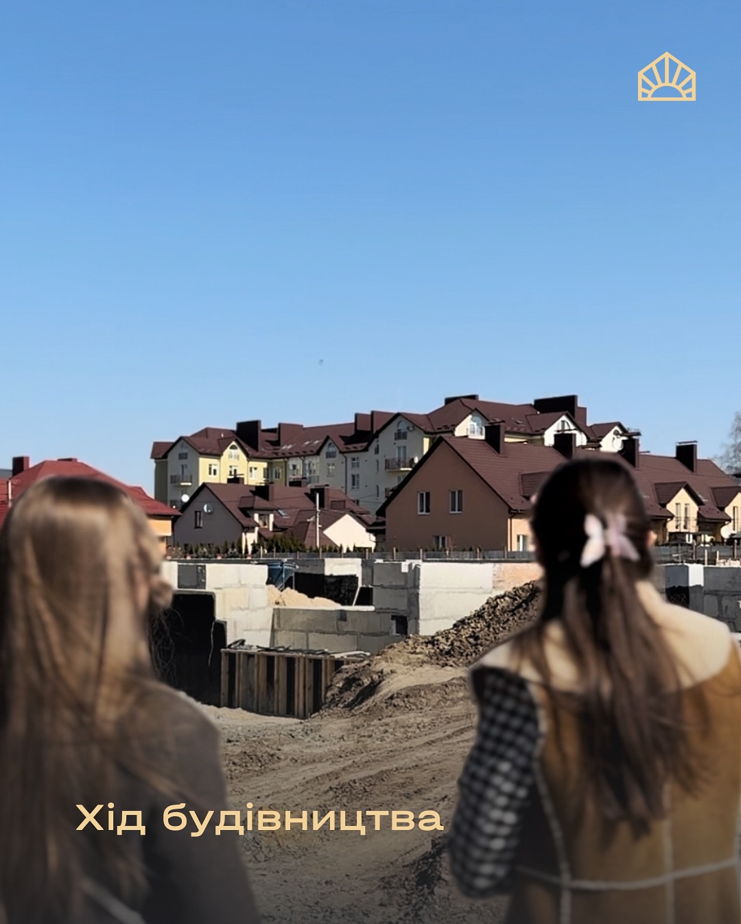 Two people with long hair stand at a construction site, watching partially built houses under a clear blue sky.