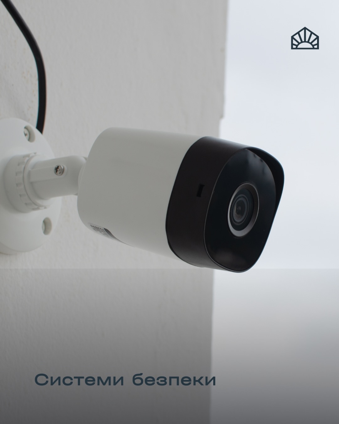 Security camera mounted on a white wall, black front facing outward, with a security logo in the top-right corner and Cyrillic caption below reading 'Системи безпеки'