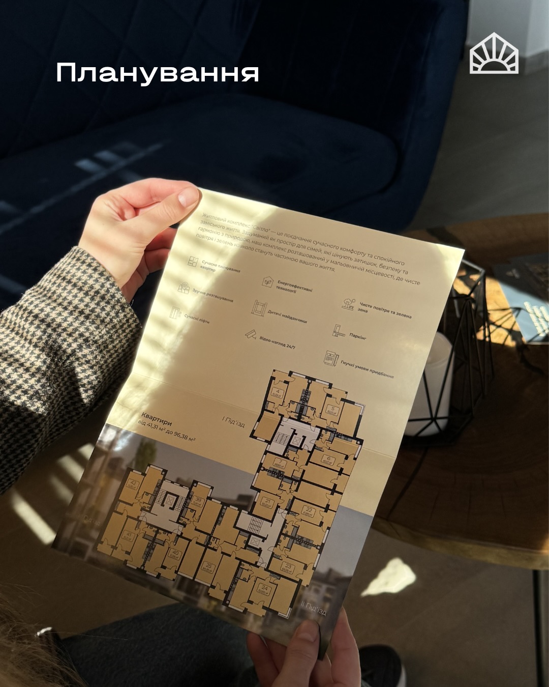 Hand holding a beige planning brochure with a floor plan layout in a living room setting, logo in corner.