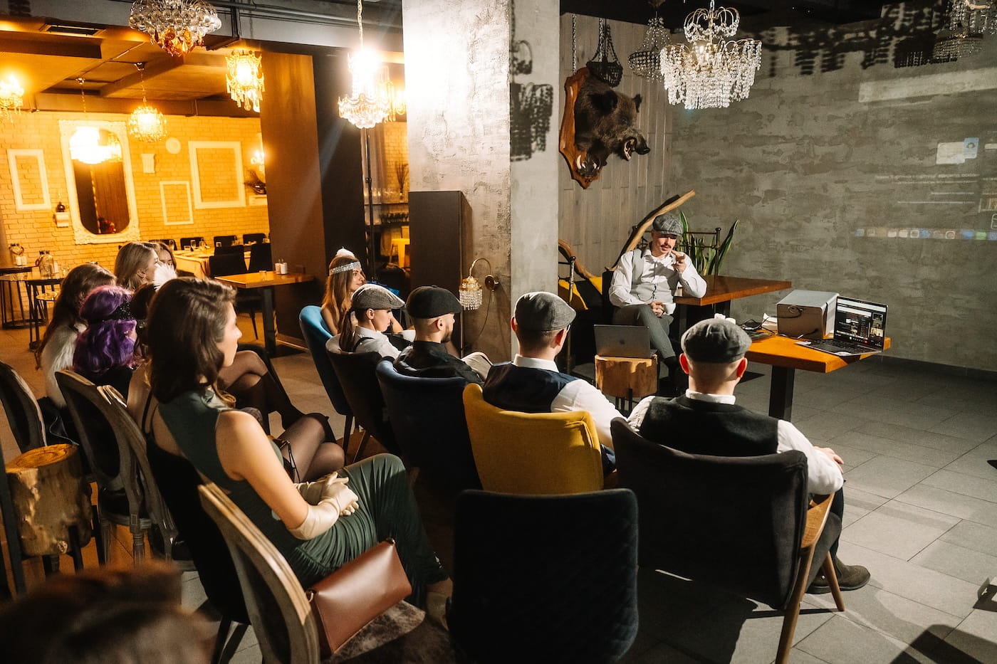 A man in vintage attire speaks to an audience seated in a cozy lounge with chandeliers and warm lighting, using a laptop and props on a table nearby.