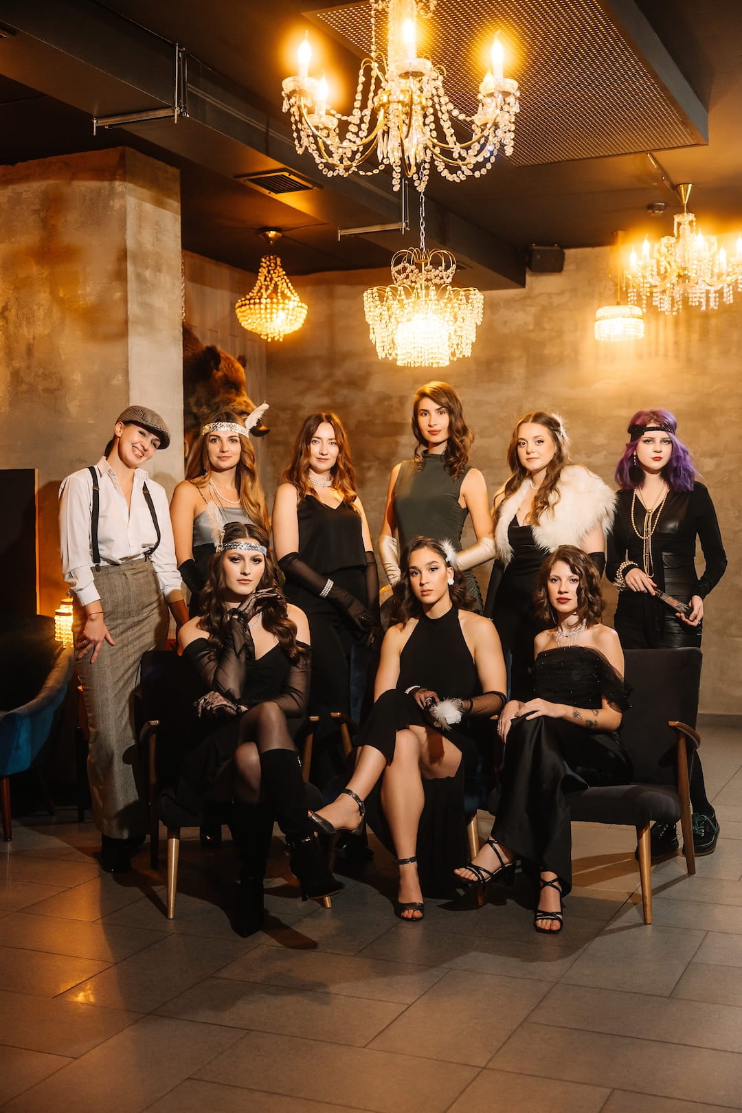 Nine women in 1920s-inspired outfits pose for a retro photoshoot under crystal chandeliers in a stylish lounge.