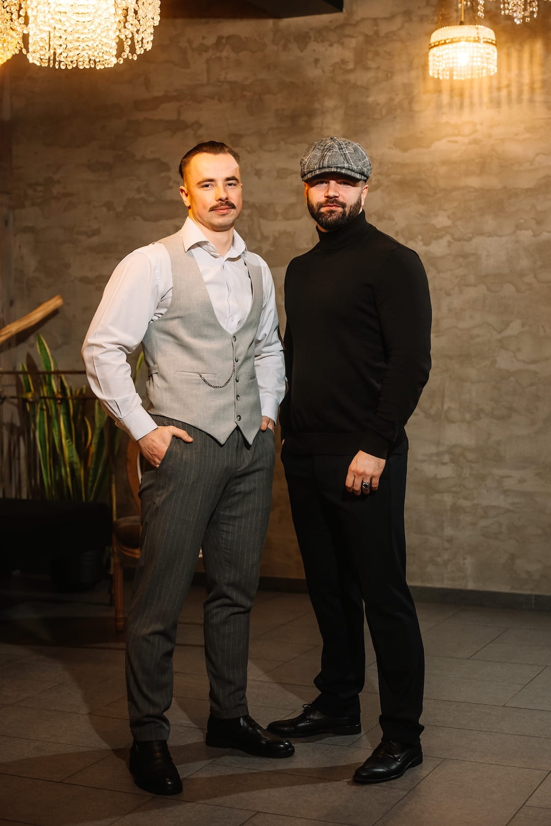 Two men pose indoors side by side: one in a light gray vest with pinstriped trousers, the other in a black turtleneck and plaid flat cap under chandelier lighting.