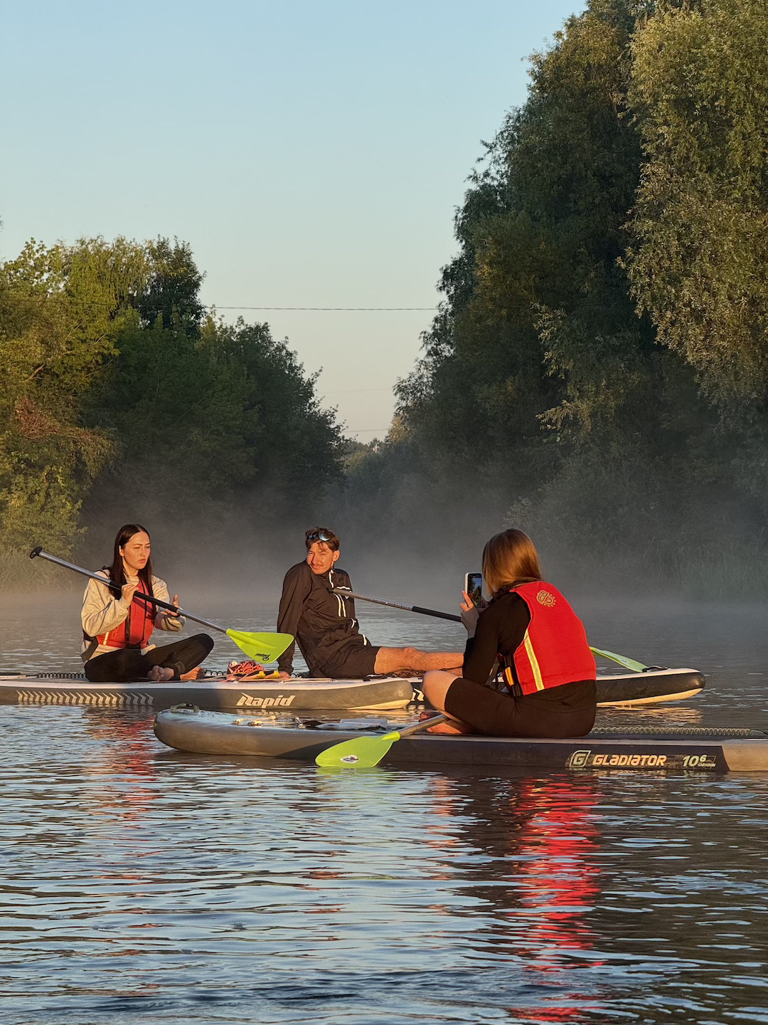 Three people on stand-up paddleboards in a misty river at dawn, one person taking a photo of others with a phone.