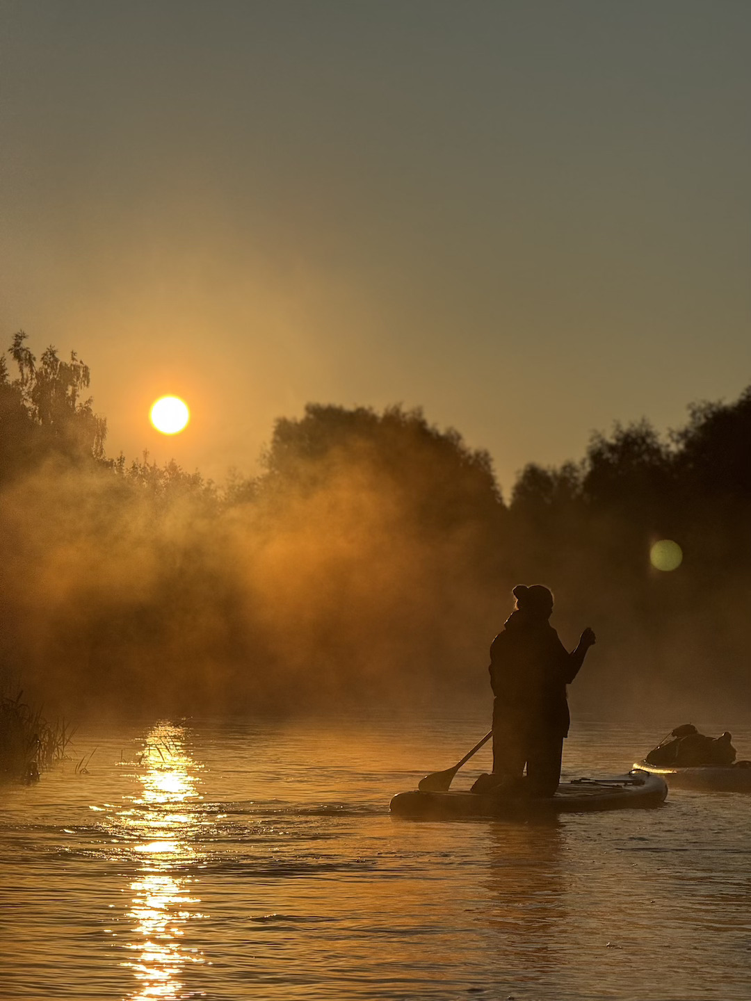 Two people stand-up paddleboarding on a misty river at sunset, silhouettes against an orange sky and reflecting light.
