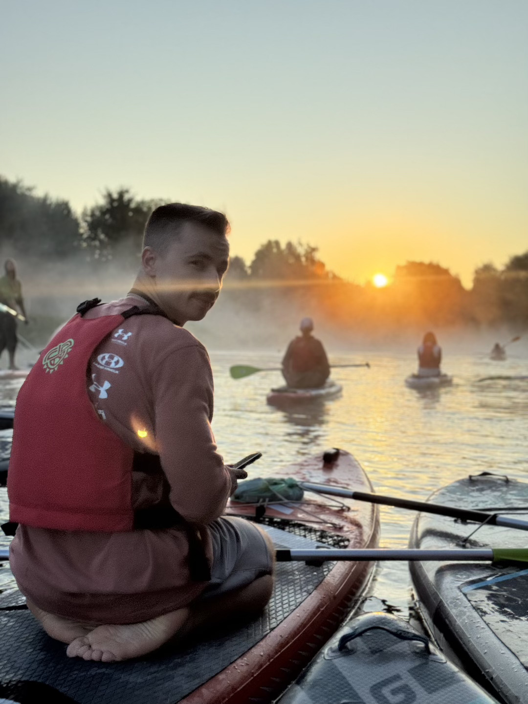 Man on a paddleboard in a red life jacket looks back at the camera as others paddle on a lake at sunset.