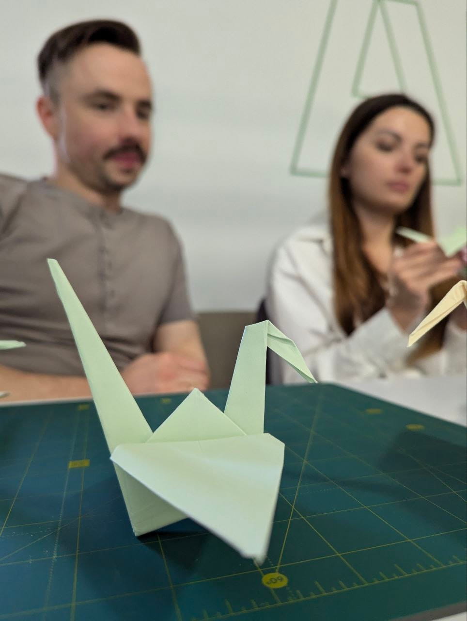 Pastel green origami crane on a grid cutting mat, with two people in soft focus in the background.