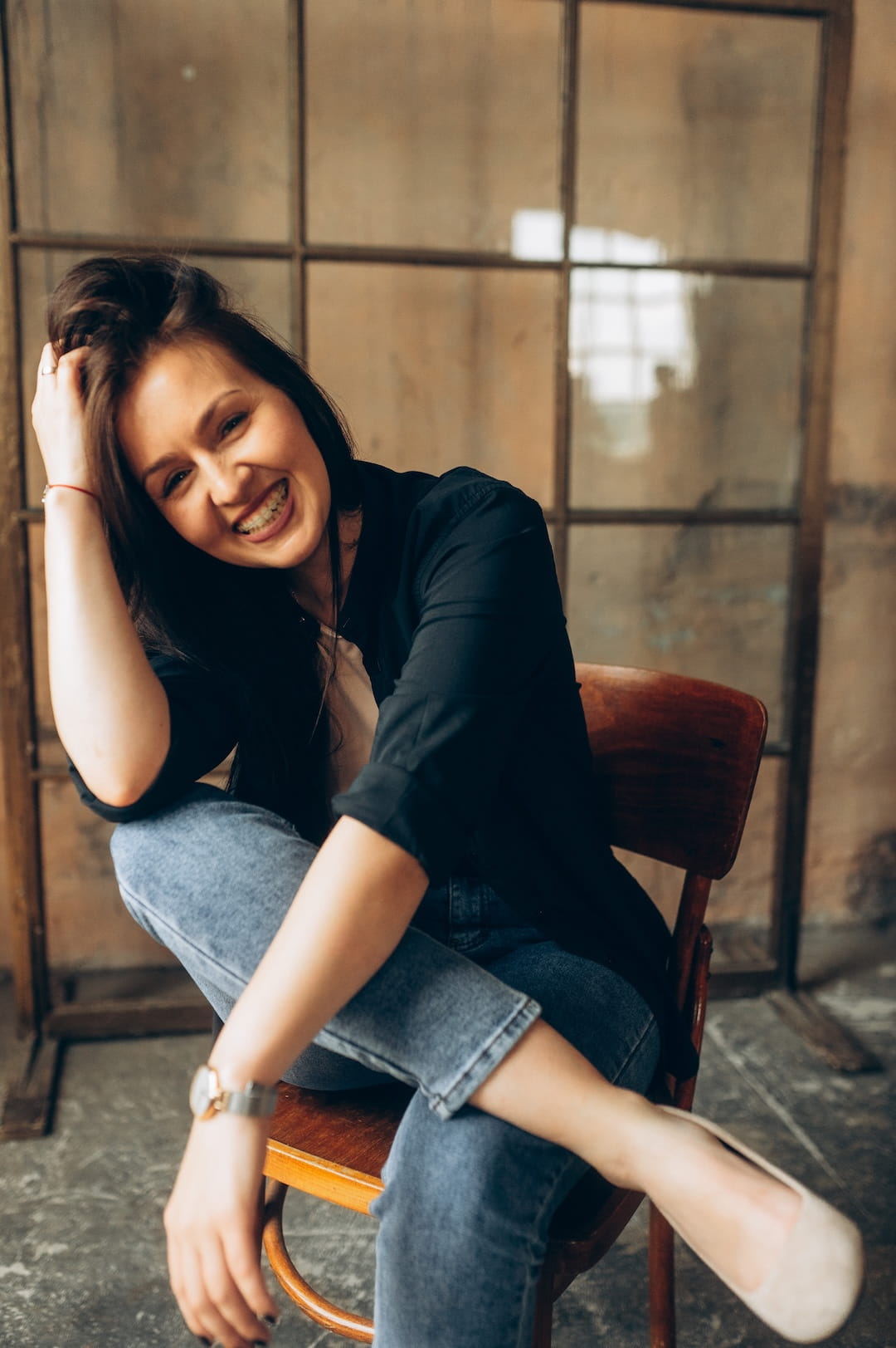 Smiling young woman sits casually on a wooden chair, one hand in her hair, wearing a black shirt and jeans in a rustic studio with large grid windows behind her.
