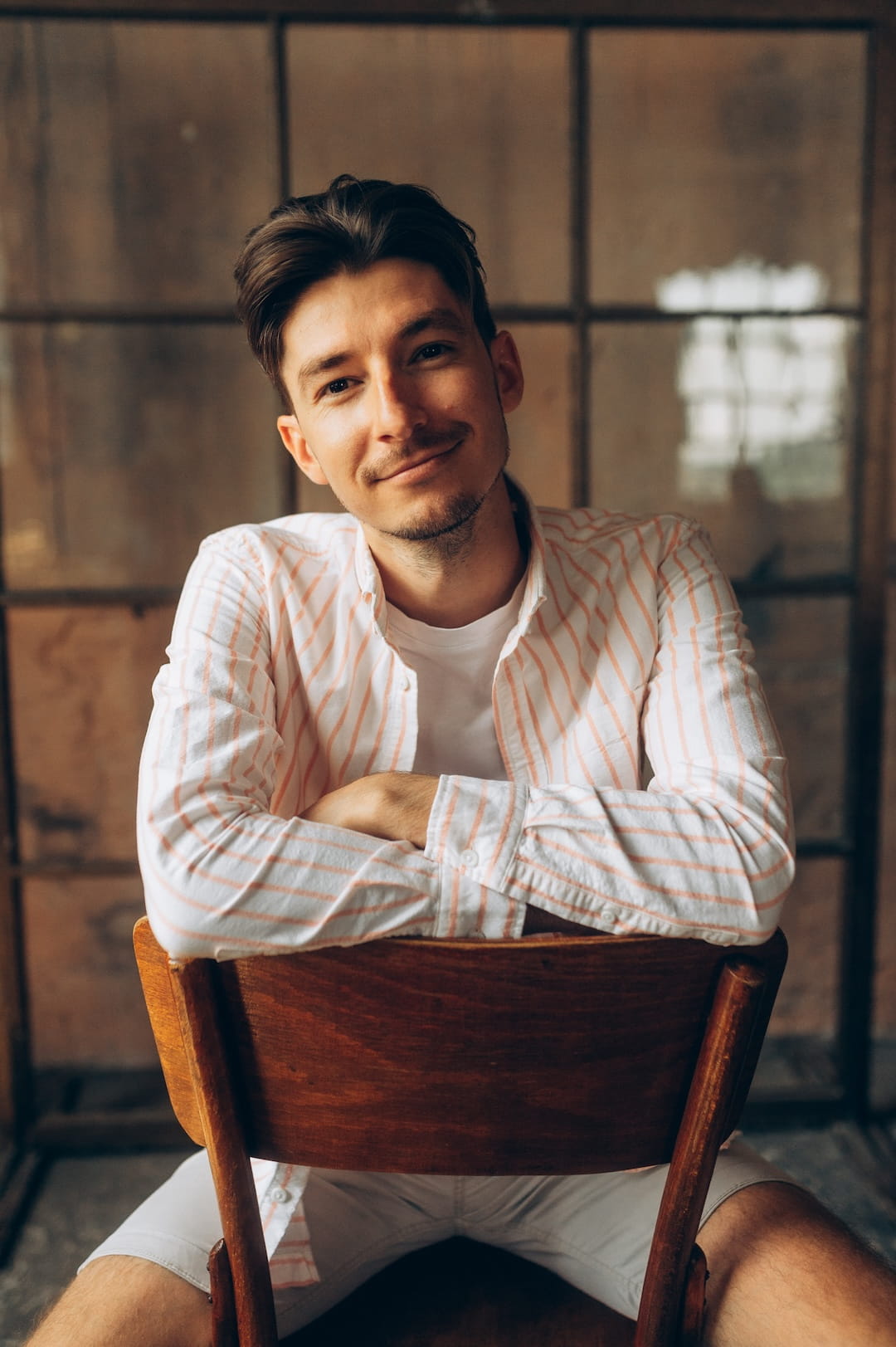 Smiling young man with brown hair sits with arms crossed on the back of a wooden chair in a warm, rustic room, wearing a white shirt with light pink stripes.