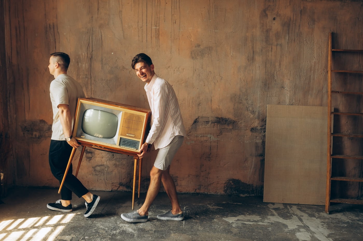 Two men carry a vintage wooden TV on a stand through a bare, rustic room; one looks back and smiles.
