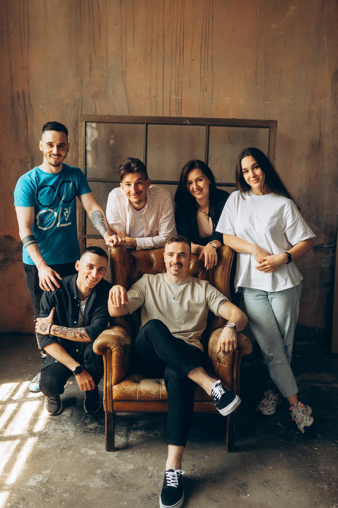 Six friends posing together in a rustic studio, with one man seated in a vintage leather chair and five others standing or kneeling around him, all smiling.