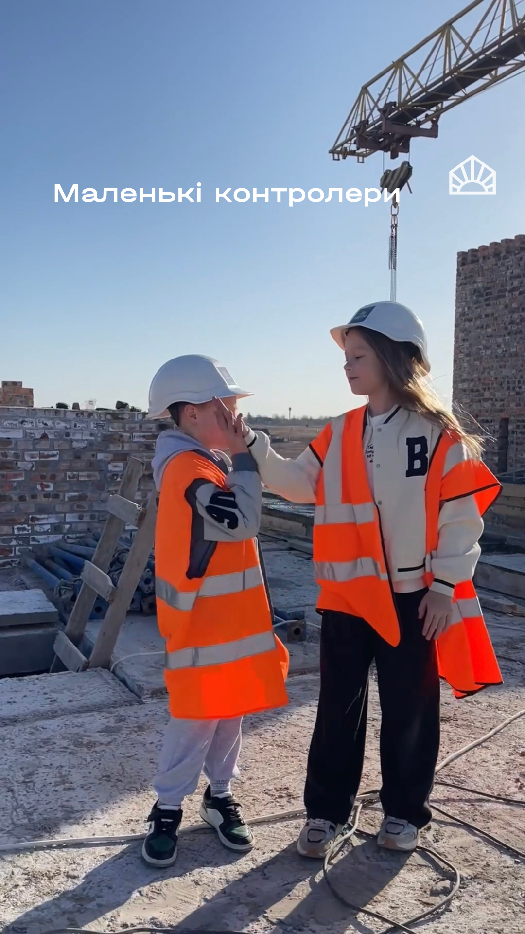 Two children in hard hats and orange safety vests on a construction site, one touching the other's cheek.