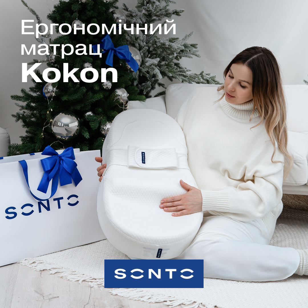 Woman in a white sweater sits on a rug, holding a white ergonomic mattress cushion beside a decorated Christmas tree and blue‑ribbon gift bag; Ukrainian marketing text for the Cocoon mattress visible.