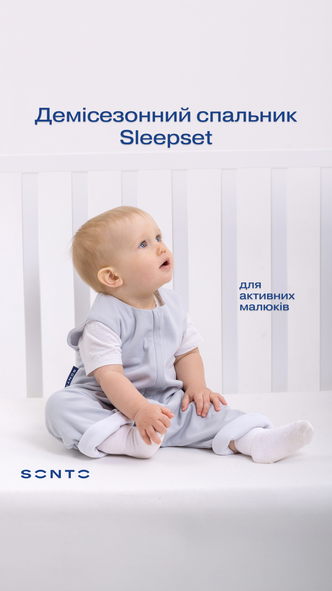 Baby in a pale blue sleepset sits on a white surface in a crib, looking upward; product photo for demi-season sleepset.