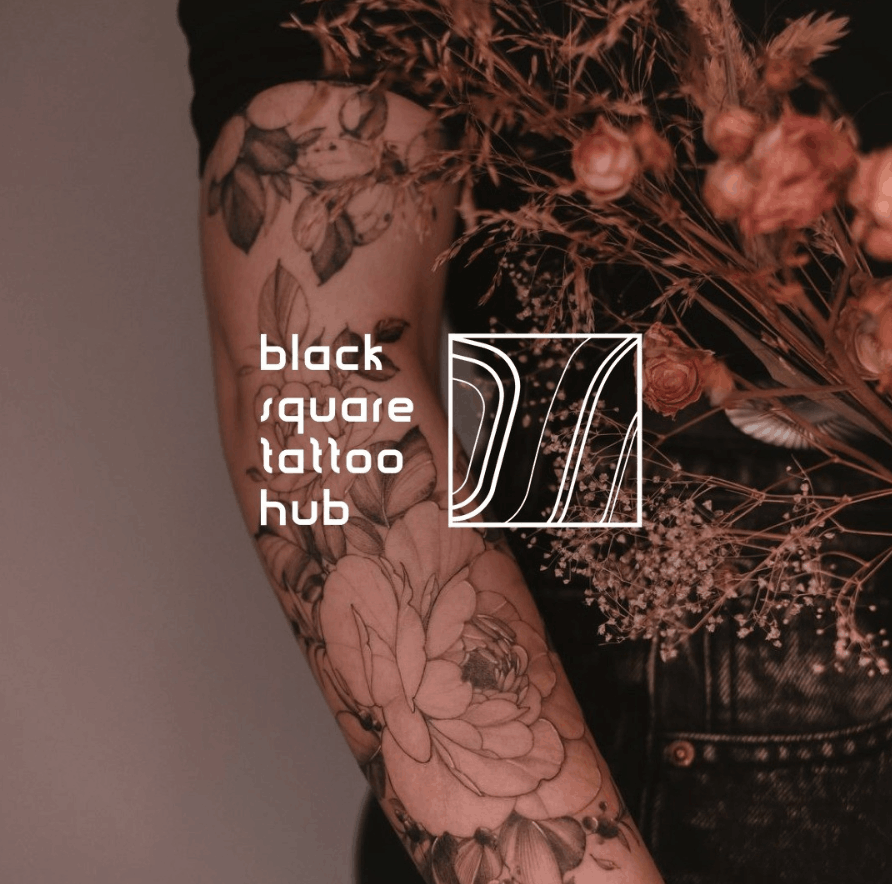 Floral tattoo on a forearm with a white logo reading 'black square tattoo hub' and a geometric square design nearby.