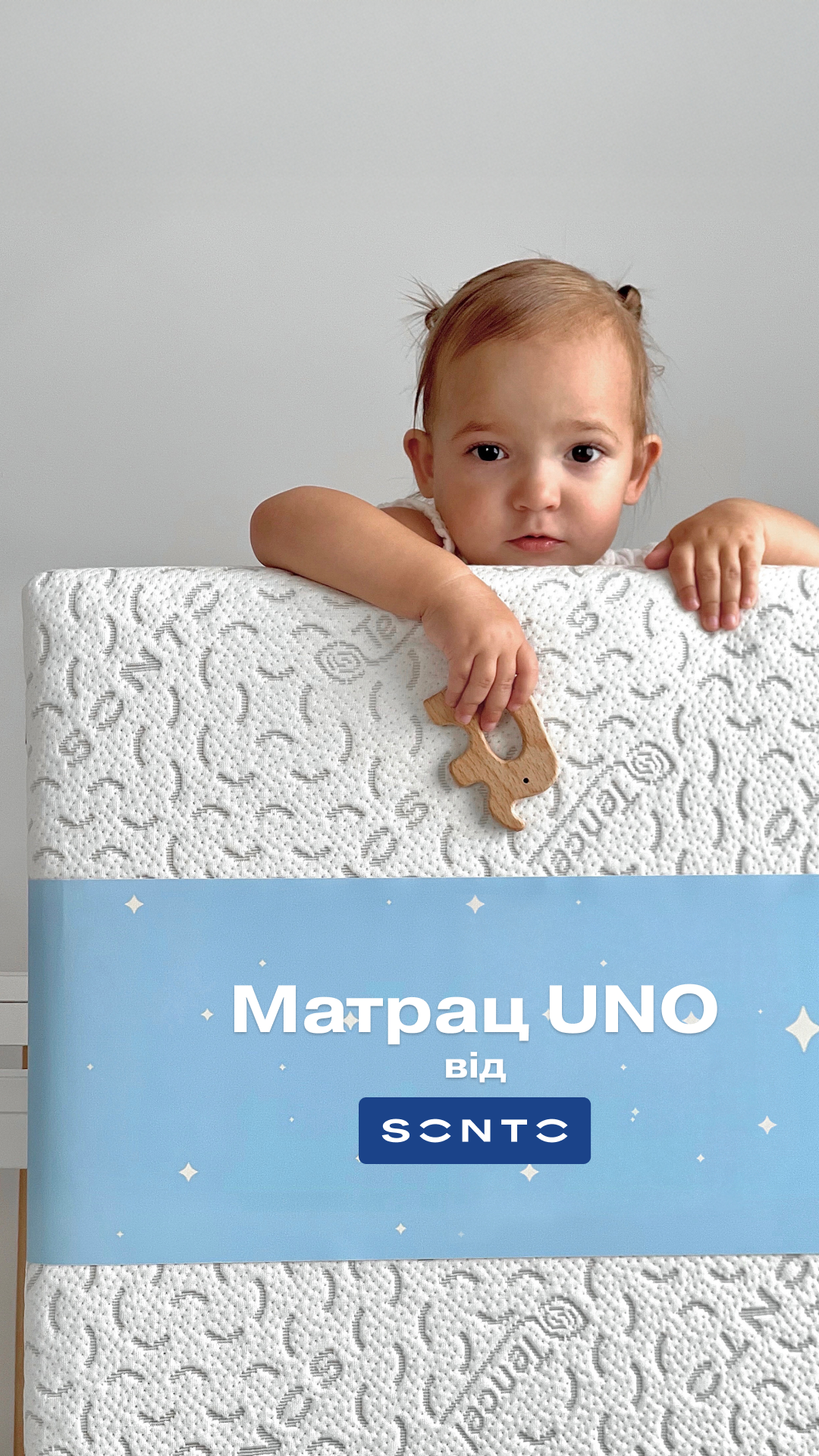 Малюк виглядає через край білого текстурного матраца з блакитним нижнім бортиком, тримаючи дерев’яну іграшку-закладку на краю.