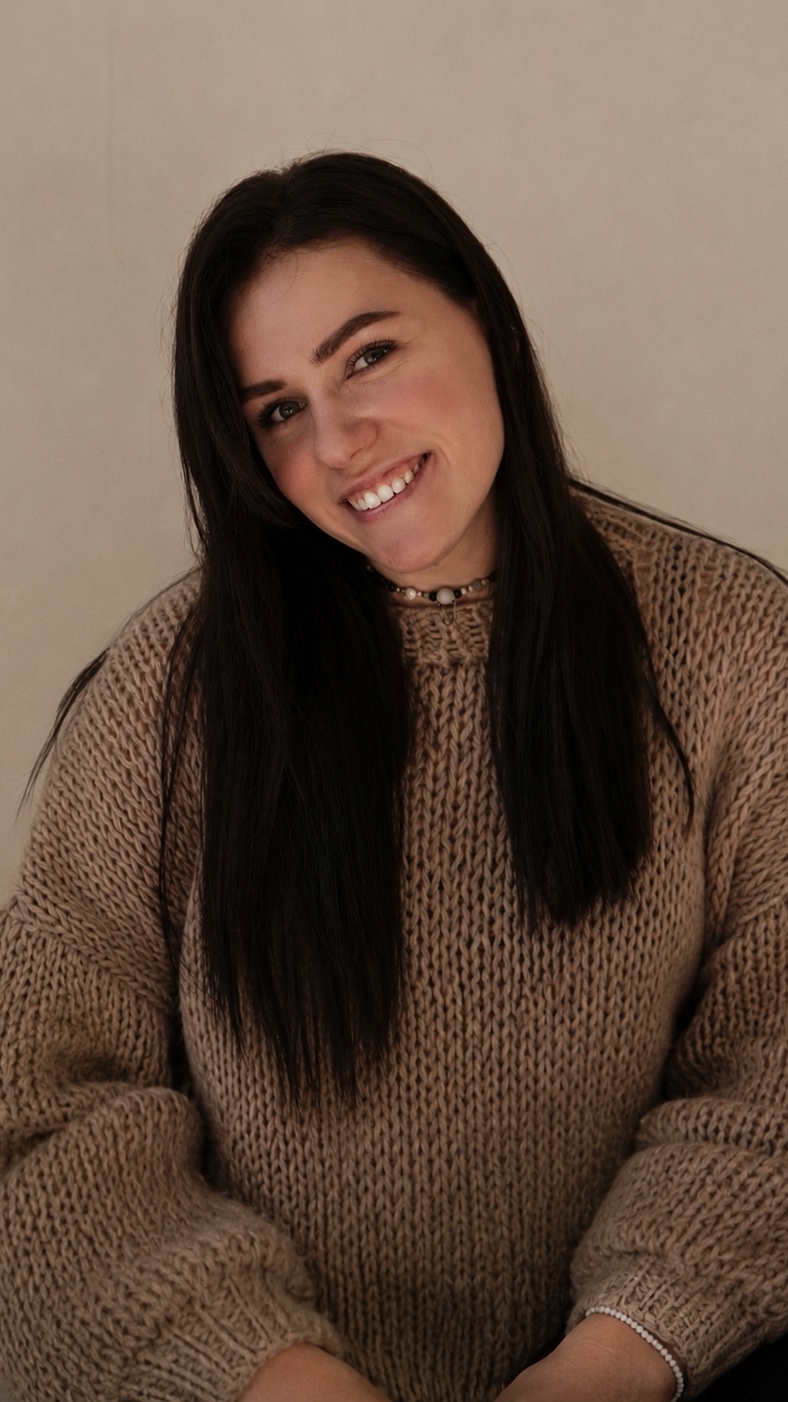 Portrait of a smiling woman with long dark hair wearing a beige chunky knit sweater against a neutral background.