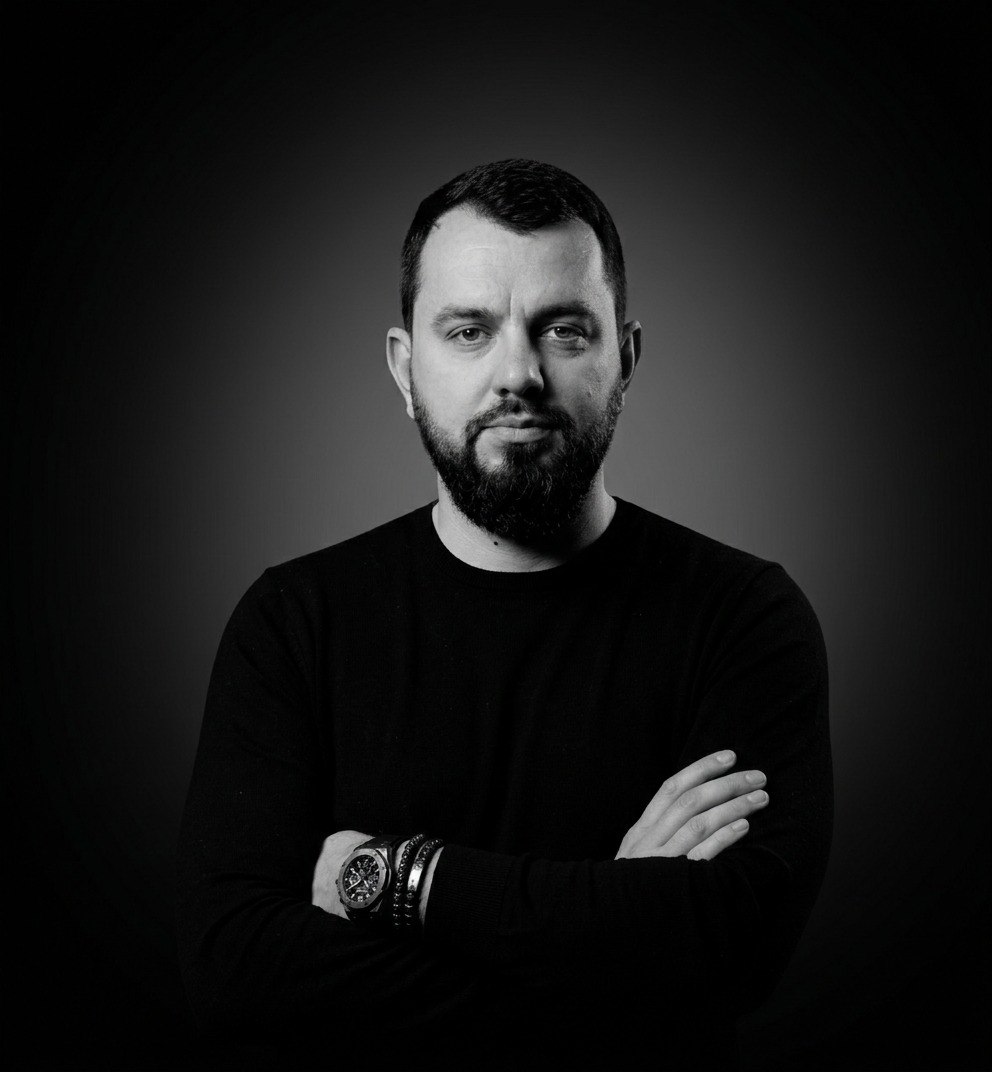 Studio black-and-white portrait of a man with a beard, arms crossed, wearing a black sweater and a wristwatch; neutral expression.
