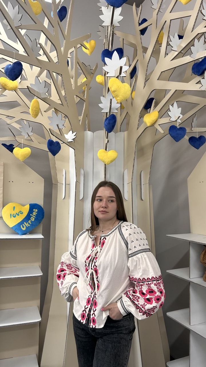 Woman in a traditional embroidered blouse stands in front of a white decorative tree with hanging blue and yellow heart ornaments; a heart reads 'Love Ukraine'.